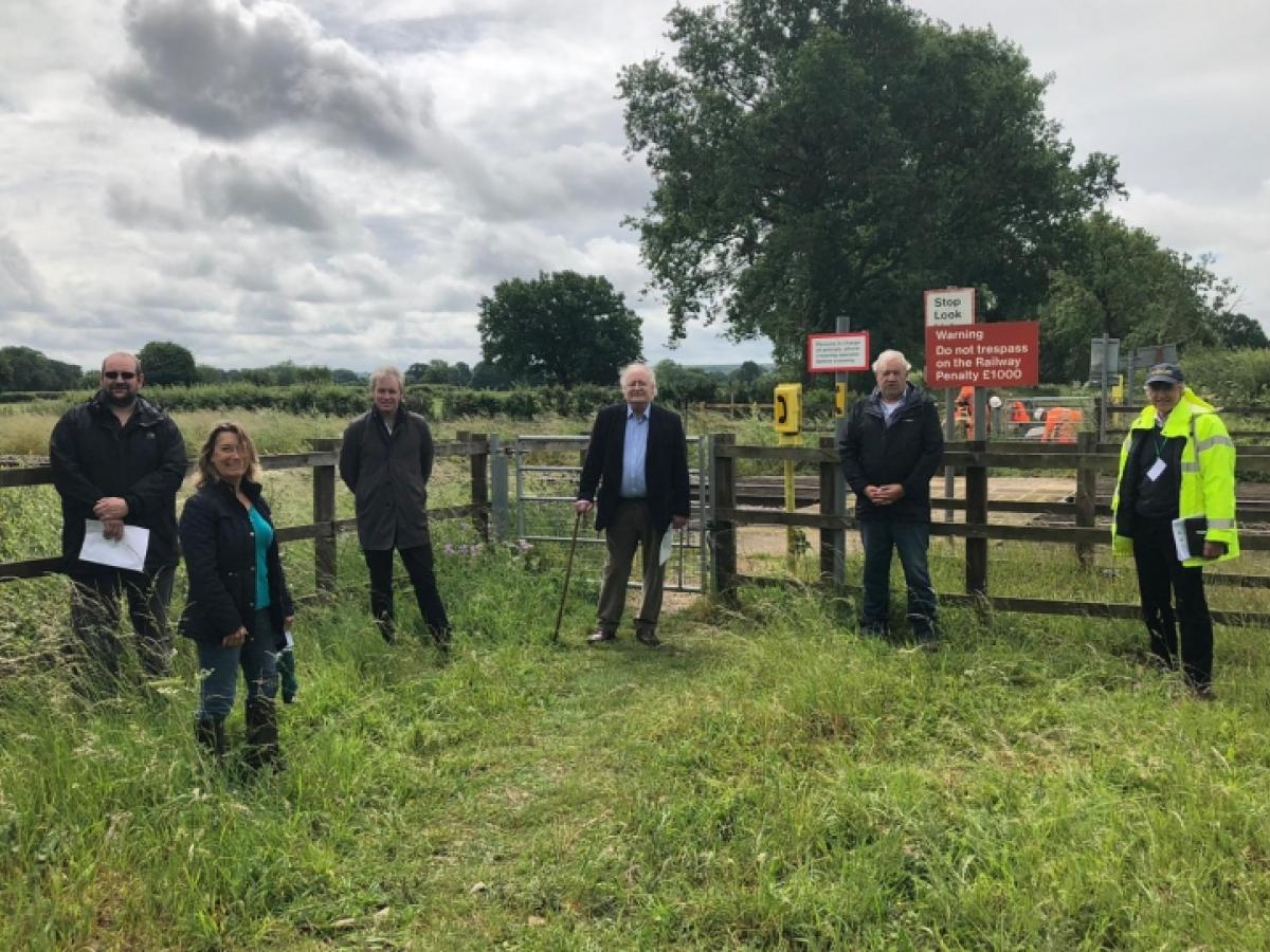 A group of people stand in a field by a fence and a railway footcrossing.