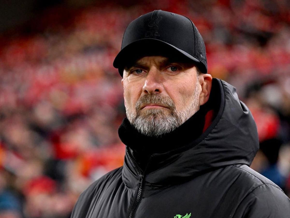 A football manager stands on the touchline and stares hard, dressed in a black coat and hat.