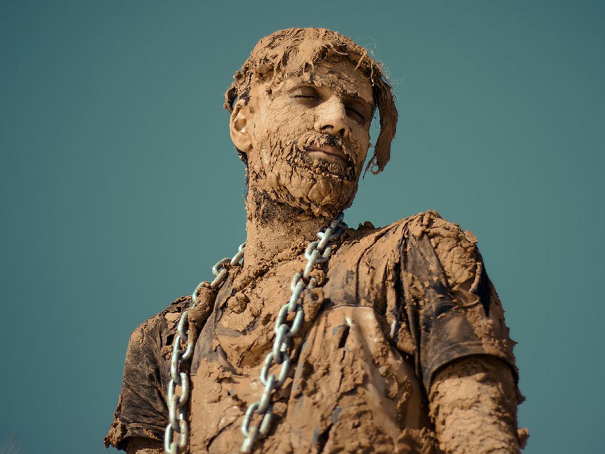 A man covered in dried and caked mud stands and looks to the side, a steel chain is draped from his shoulders.