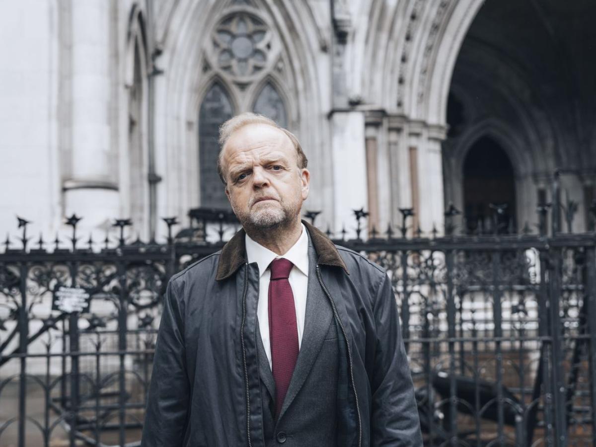 A man, dressed in a suit and anarak, stands in front of a law court.