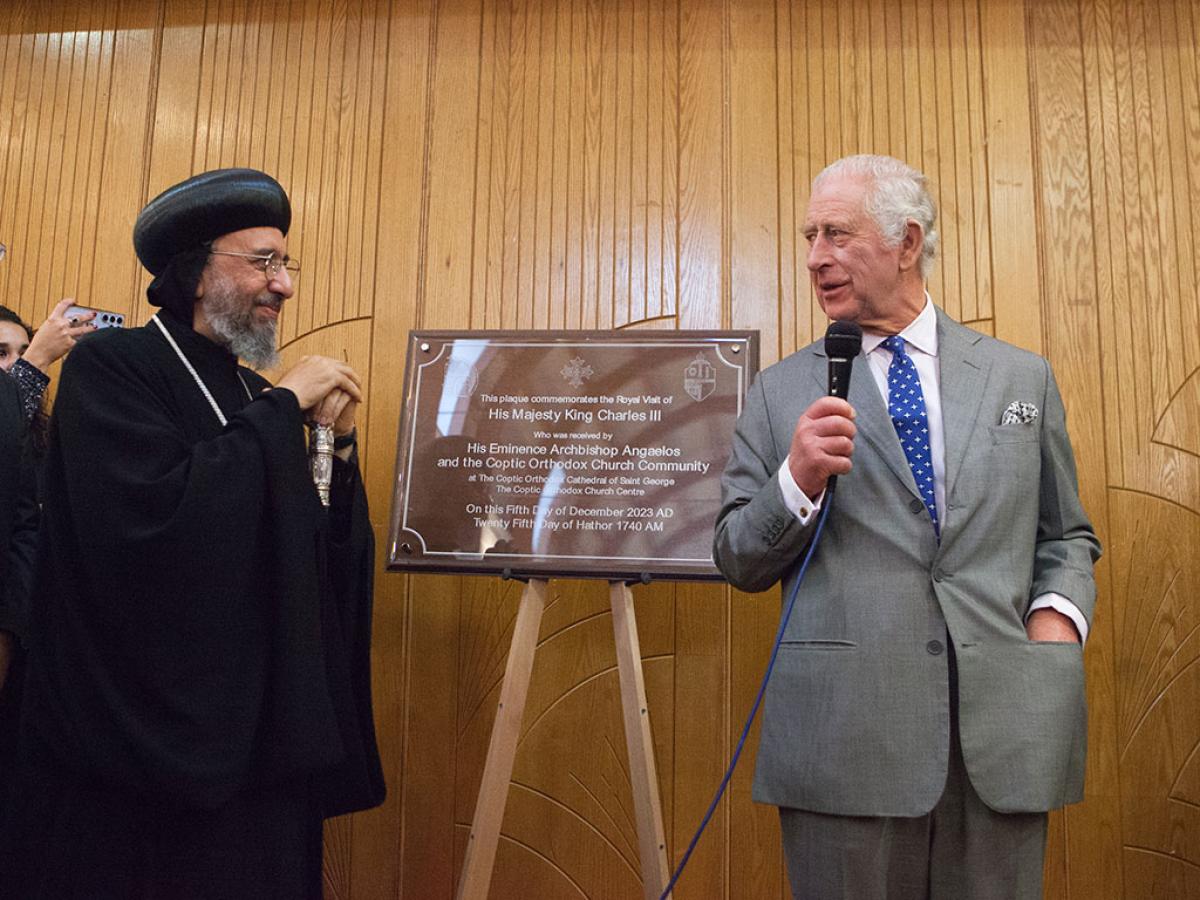 An archbishop wearing a black hat and robes stands next to a new building's plaque, while King Charles, wearing a suit stands the other side holding a mic.