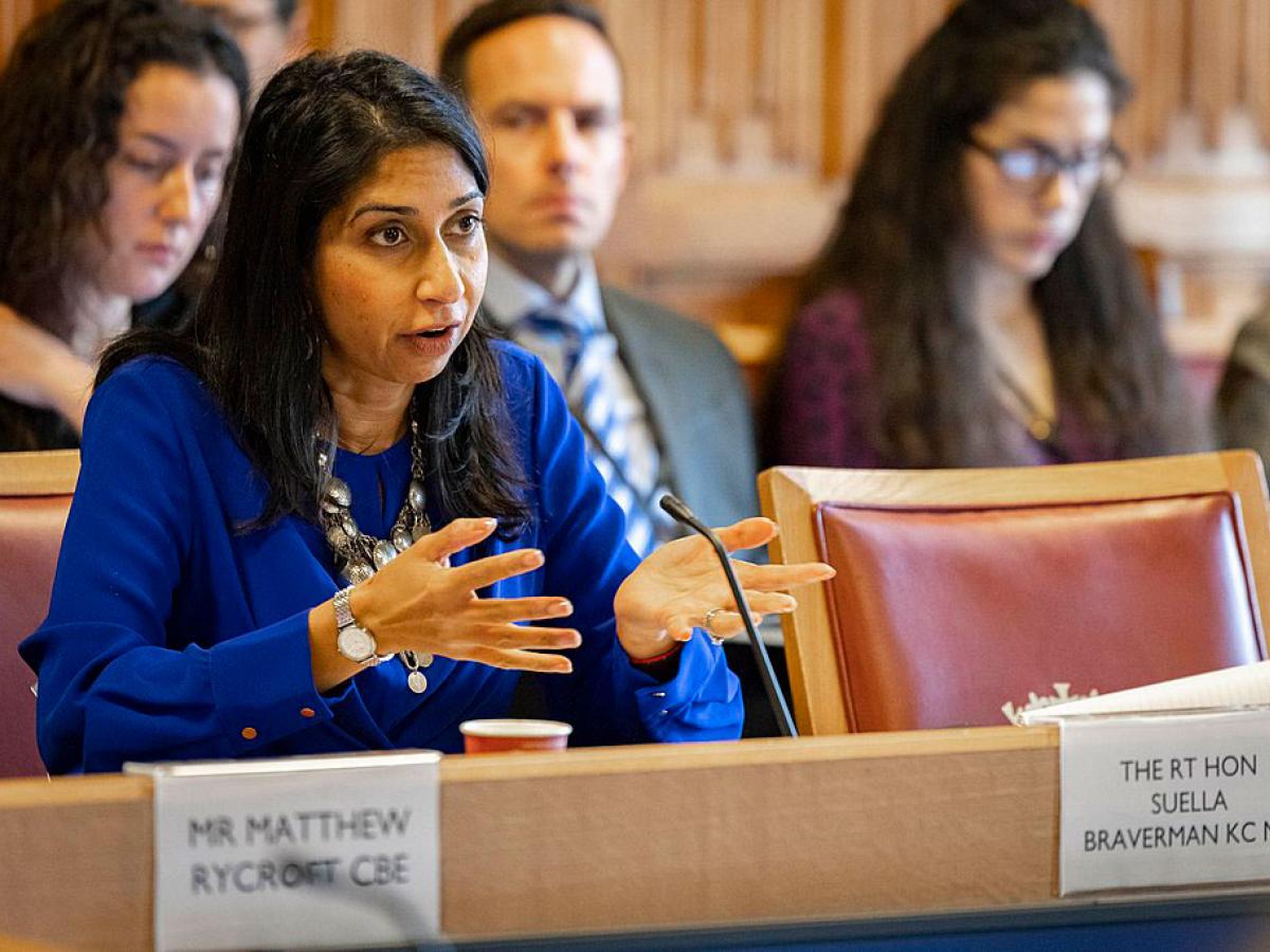 A woman dressed in a blue suit sits at a table talking and gesticulating with her hands.