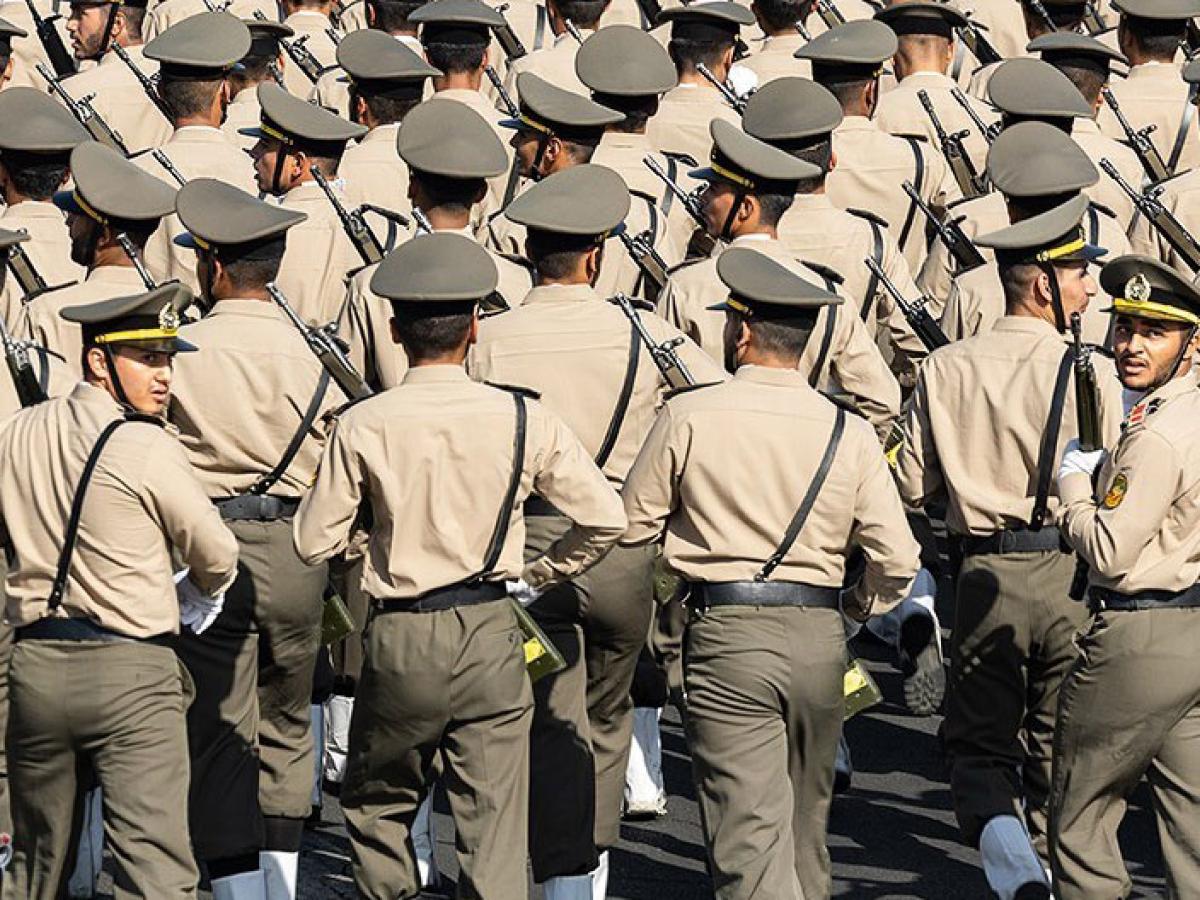 Rows of soldiers march away from the camera, two in the back row turn their heads back.