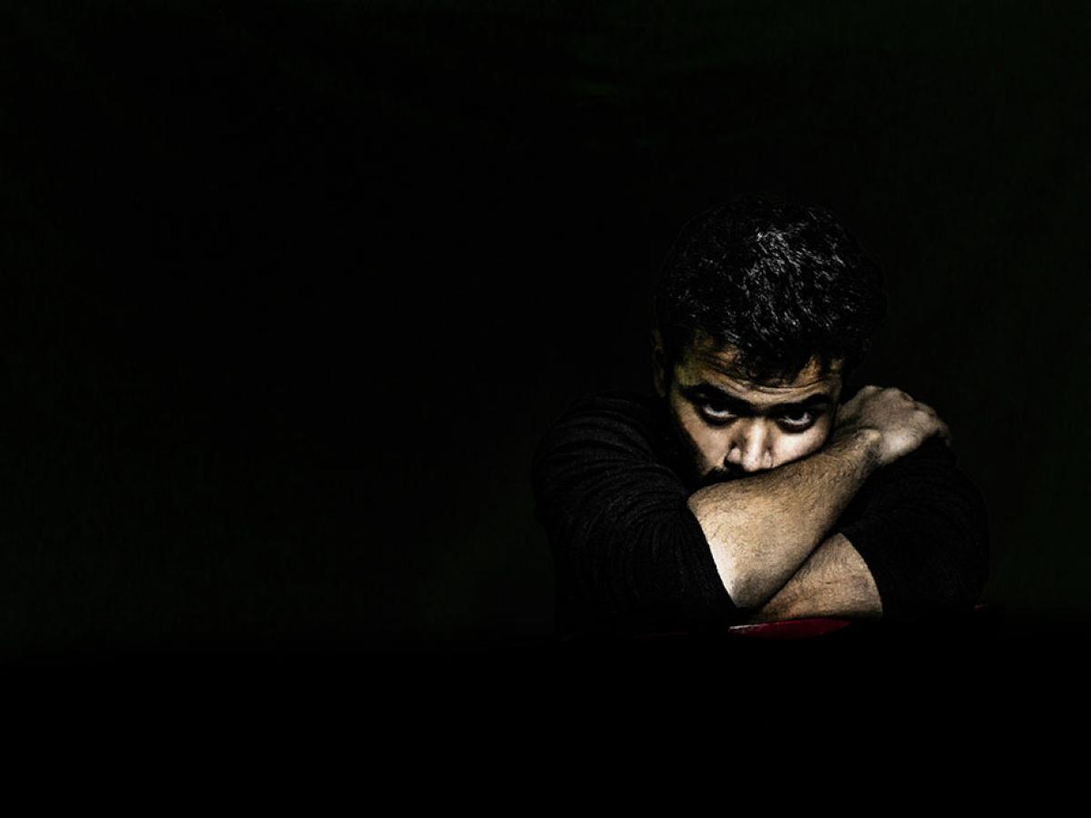 In a darkened room, a man's angry face is lit as he rests on arms folded tightly around it.