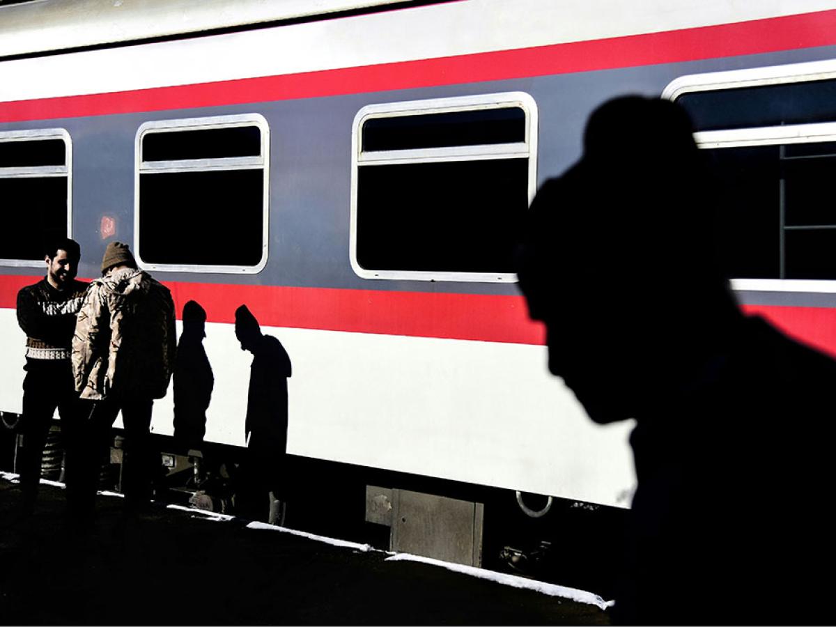 On a sun-lit railway platform, two men talk casting shadows on a waiting red and white train. Another shadow beside them is that of a man silouheted in the foreground