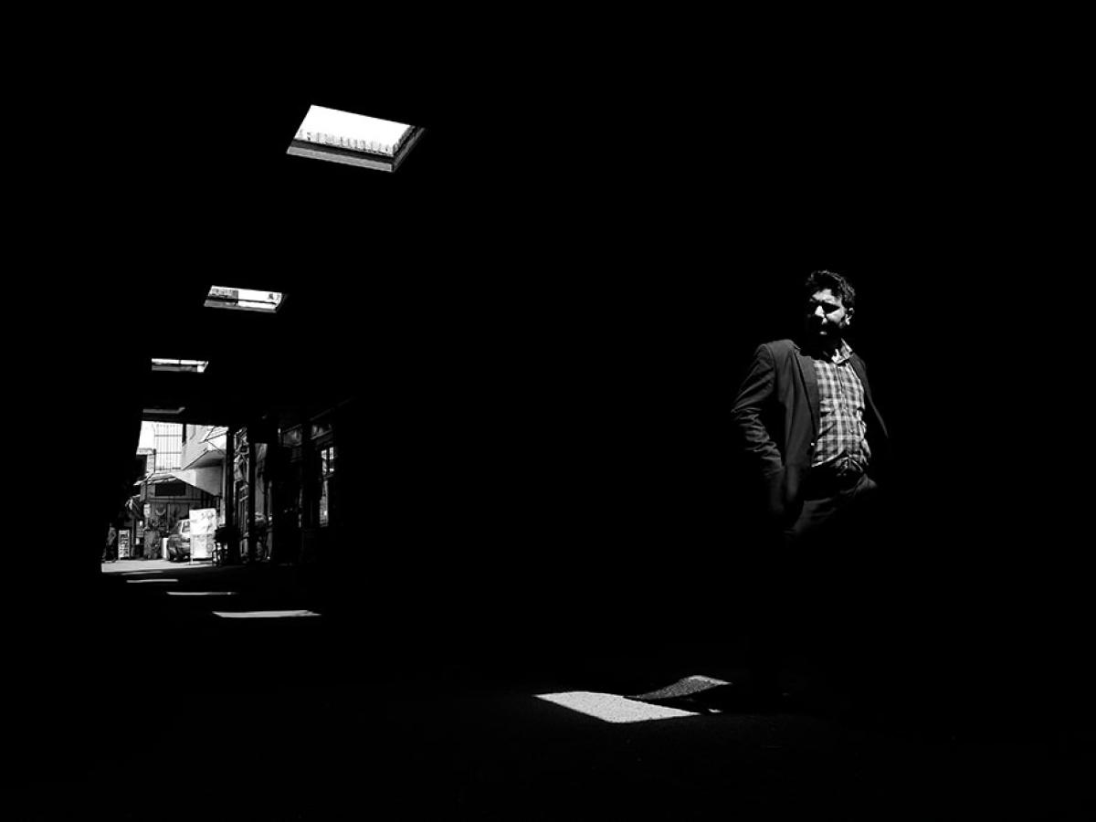 A man walks through a dark alley, looking to one side, illuminated only by roof lights.