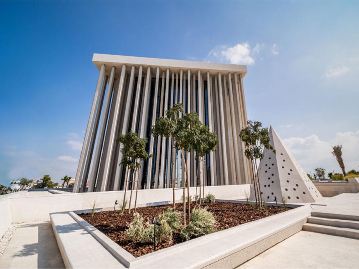 A large, modern cubiod building with a series of thin external pillars is seen from low down against a sunny blue sky 