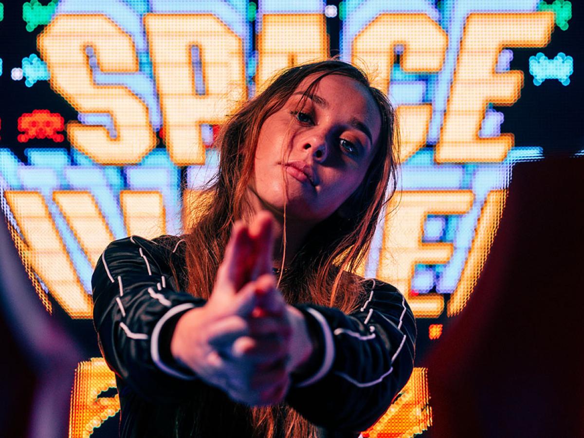 A woman stand in front of a large video screen displaying the Space Invaders title, hold her hands out in front of her.