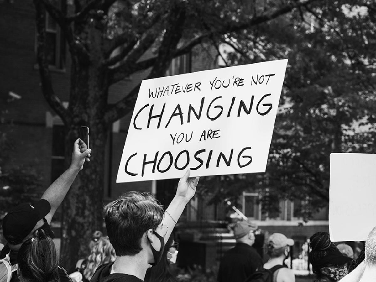A protest placard is held above a march, reading 'If you don't change it, you choose it'.