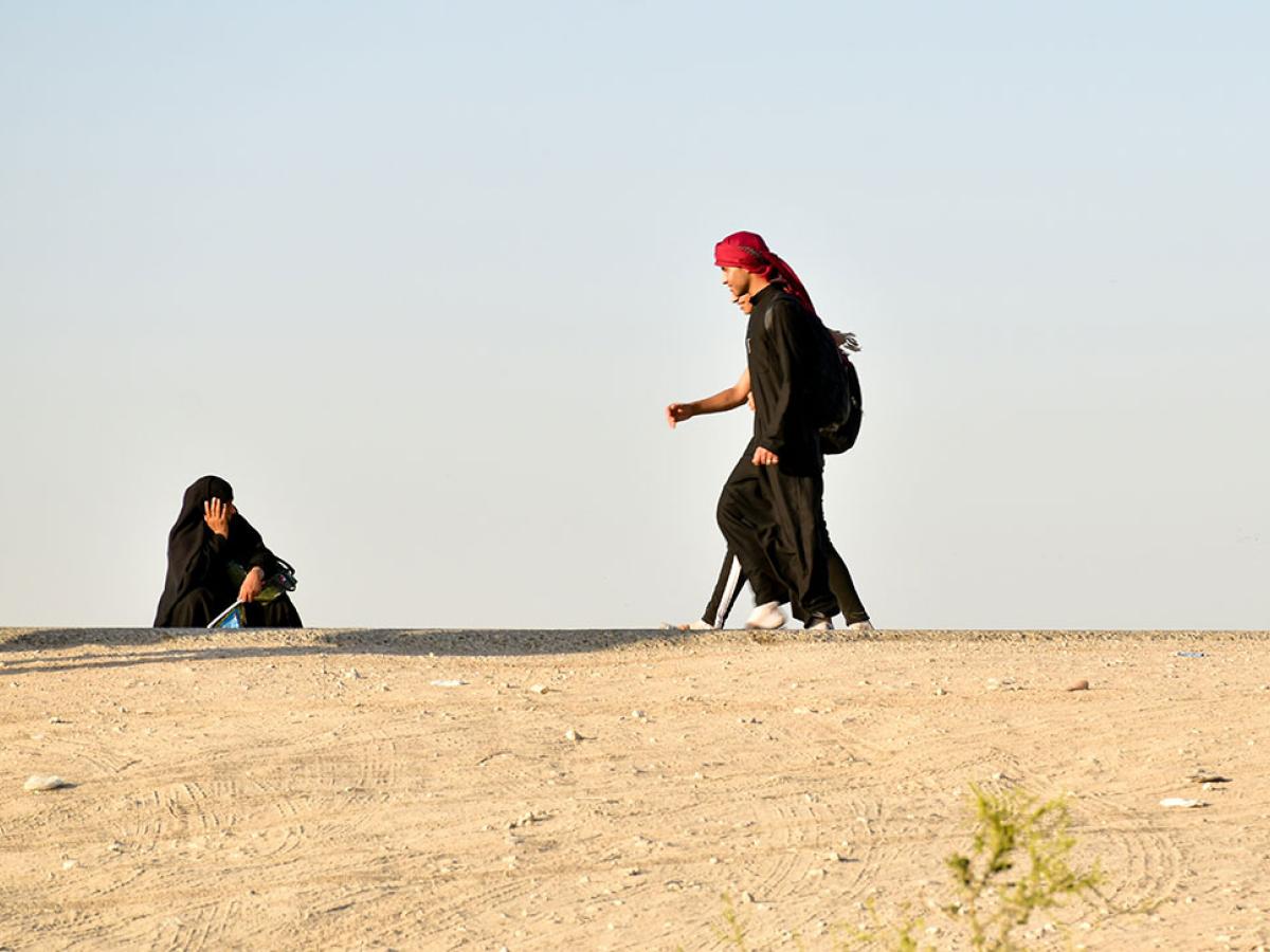 A black clad pilgrim squats and holds his head on the side of a desert road as other pilgrims walk along.