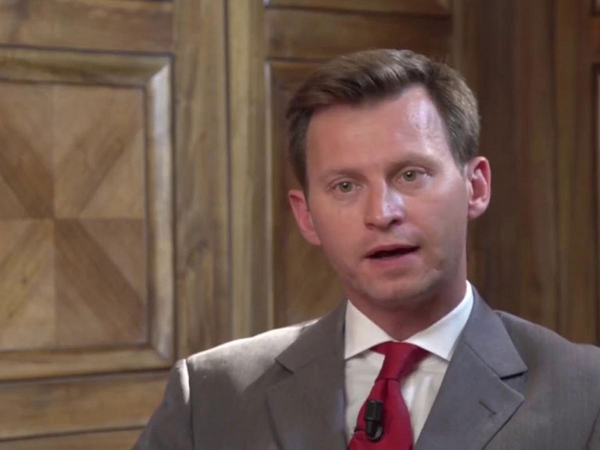 A man talks animatedly looking at the camera while sitting against a wood panelled wall.