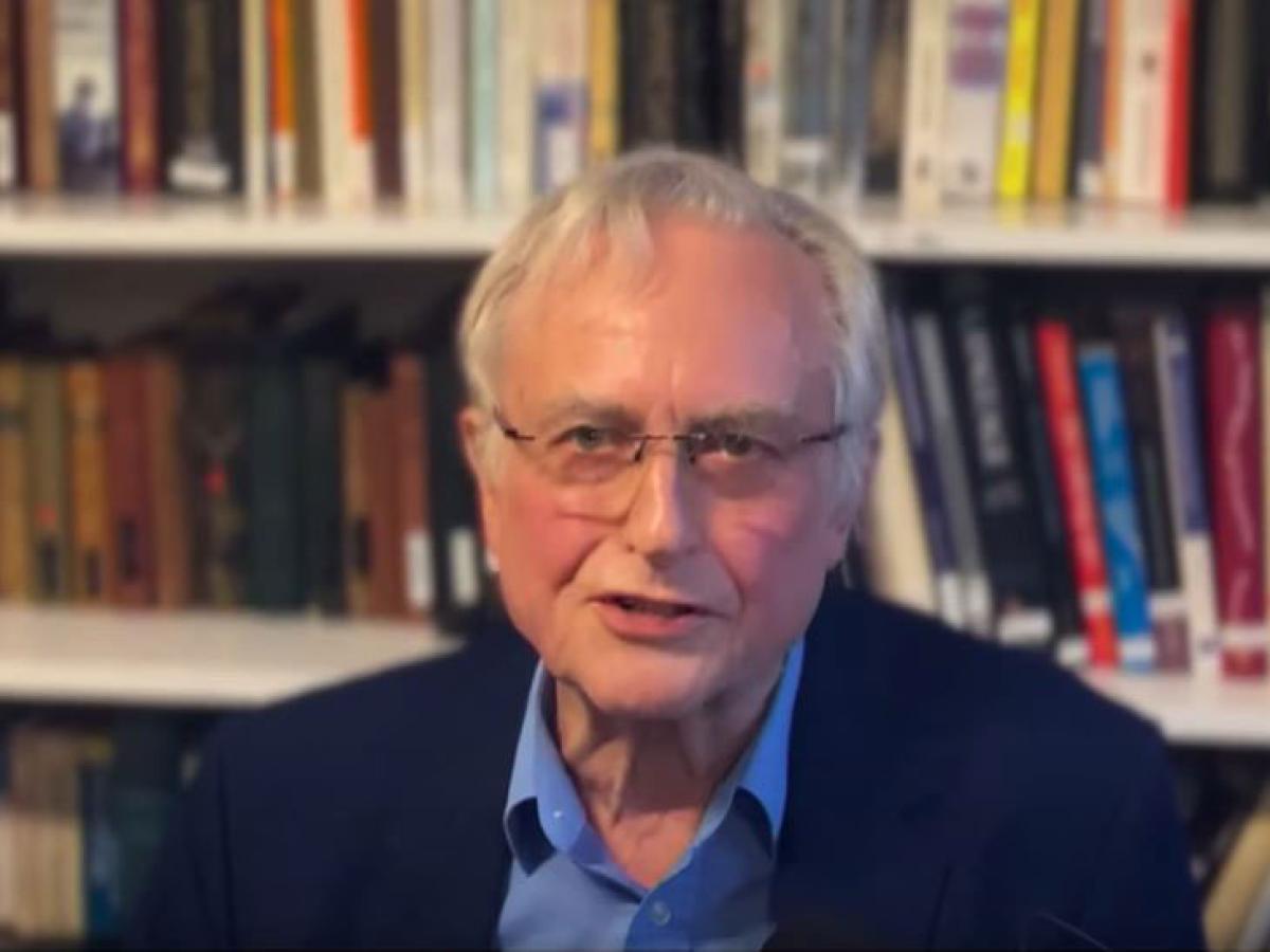 A man sits and speaks, against a background of a bookcase.