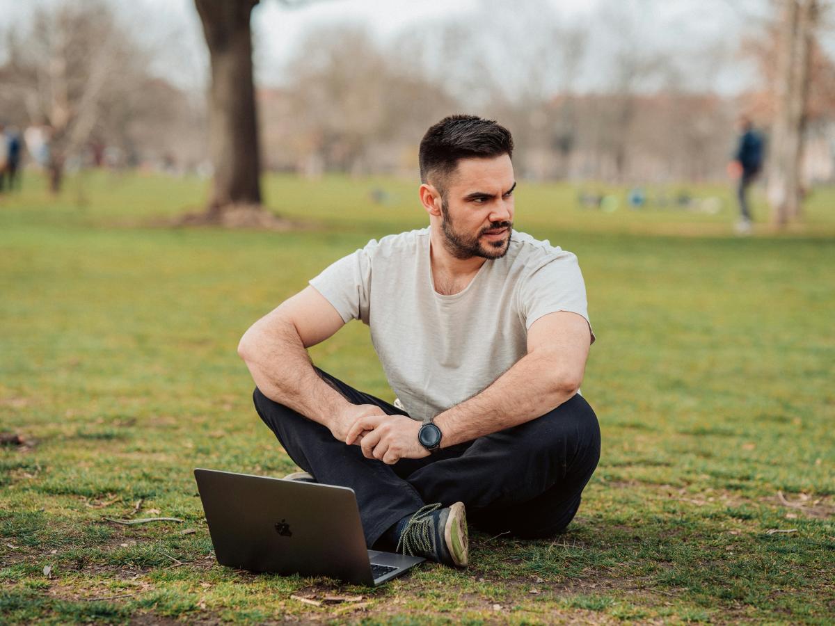 A man sits cross legged in a park with a laptop on the grass in front of him. He looks to one side.