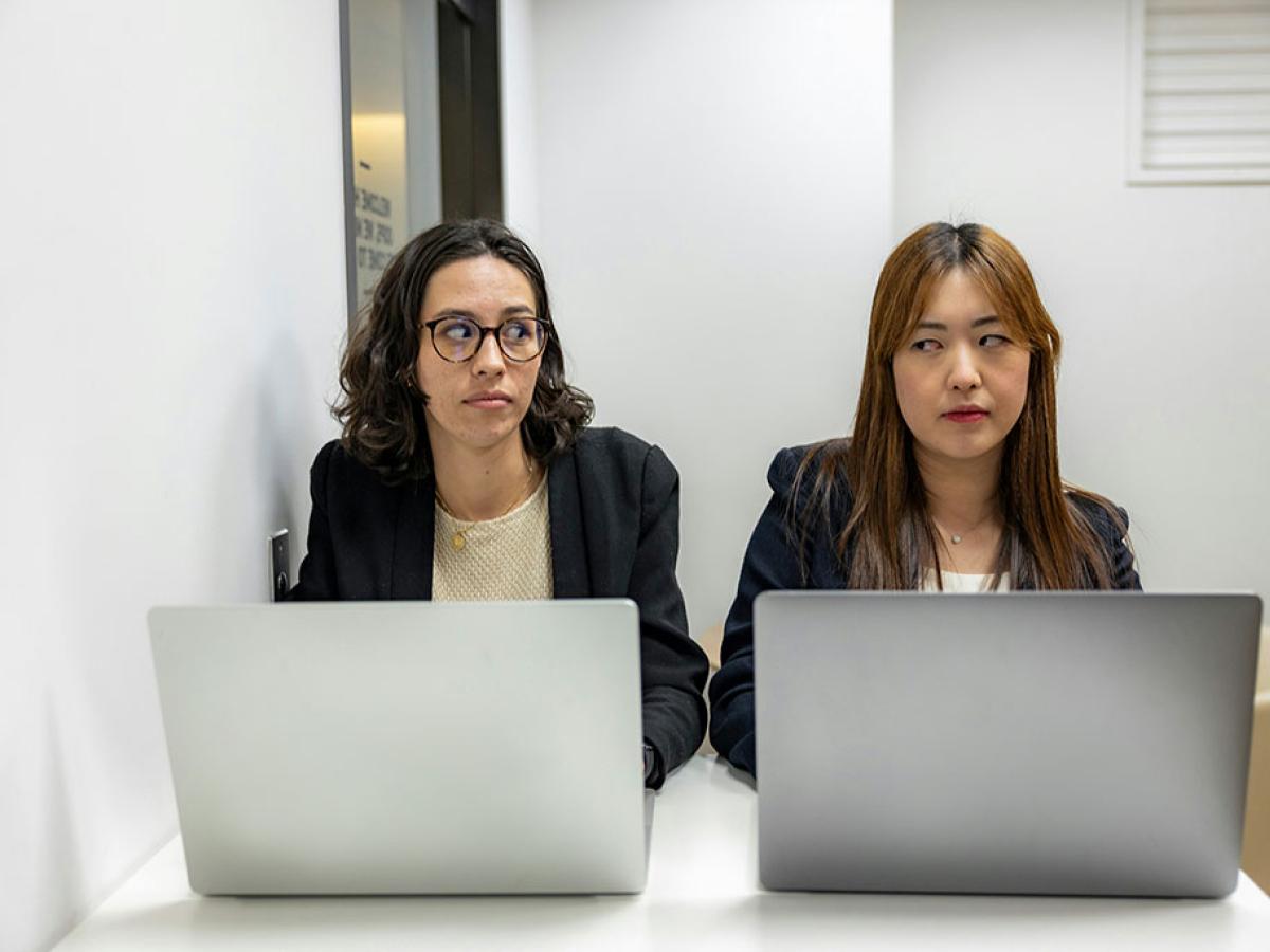 Two people sit beside each other working on laptops, One looks askance to the other who frowns.