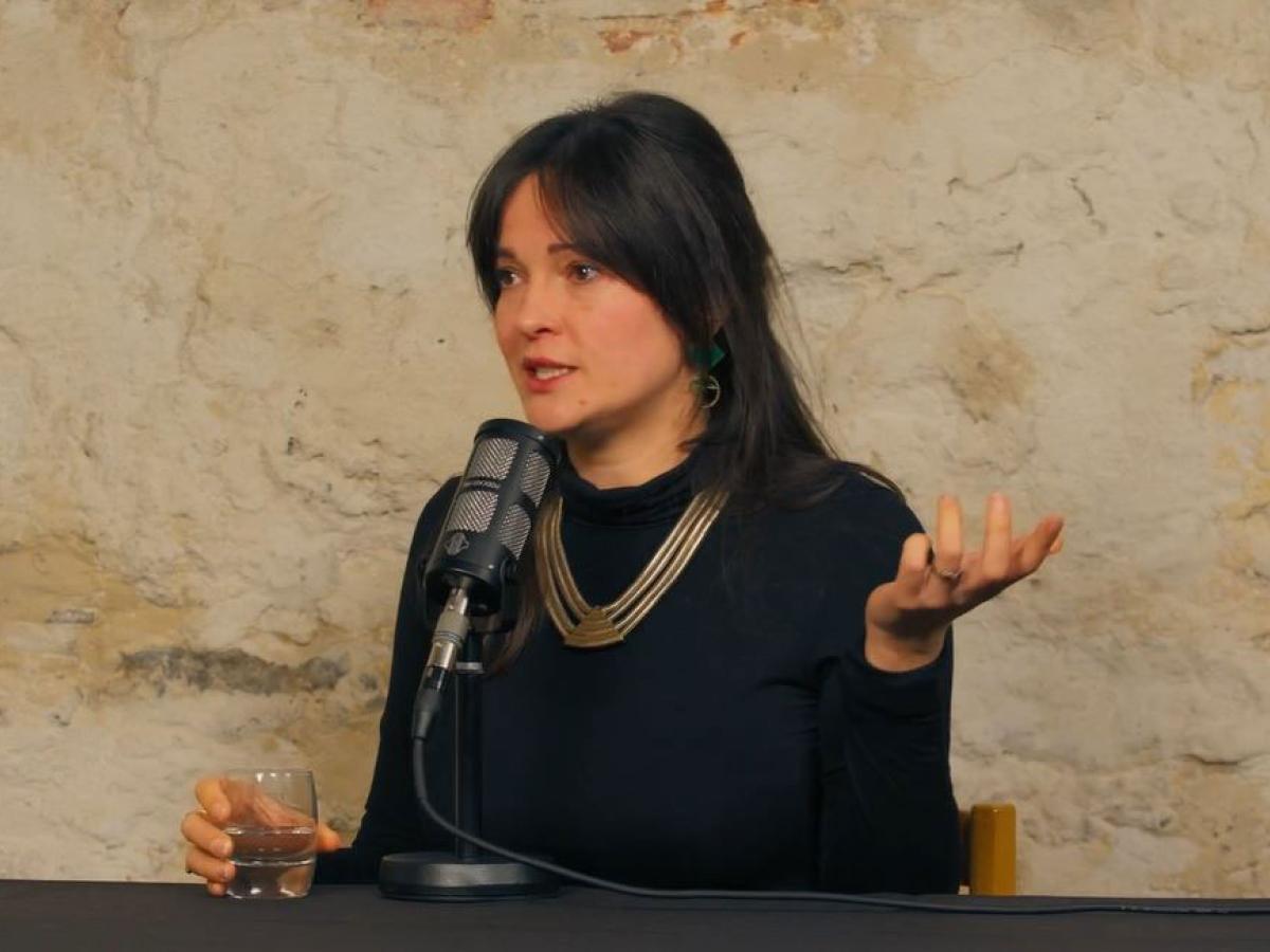 A woman sits a a table with a microphone in front of her, she is talking and gestures with one raise hand upward.
