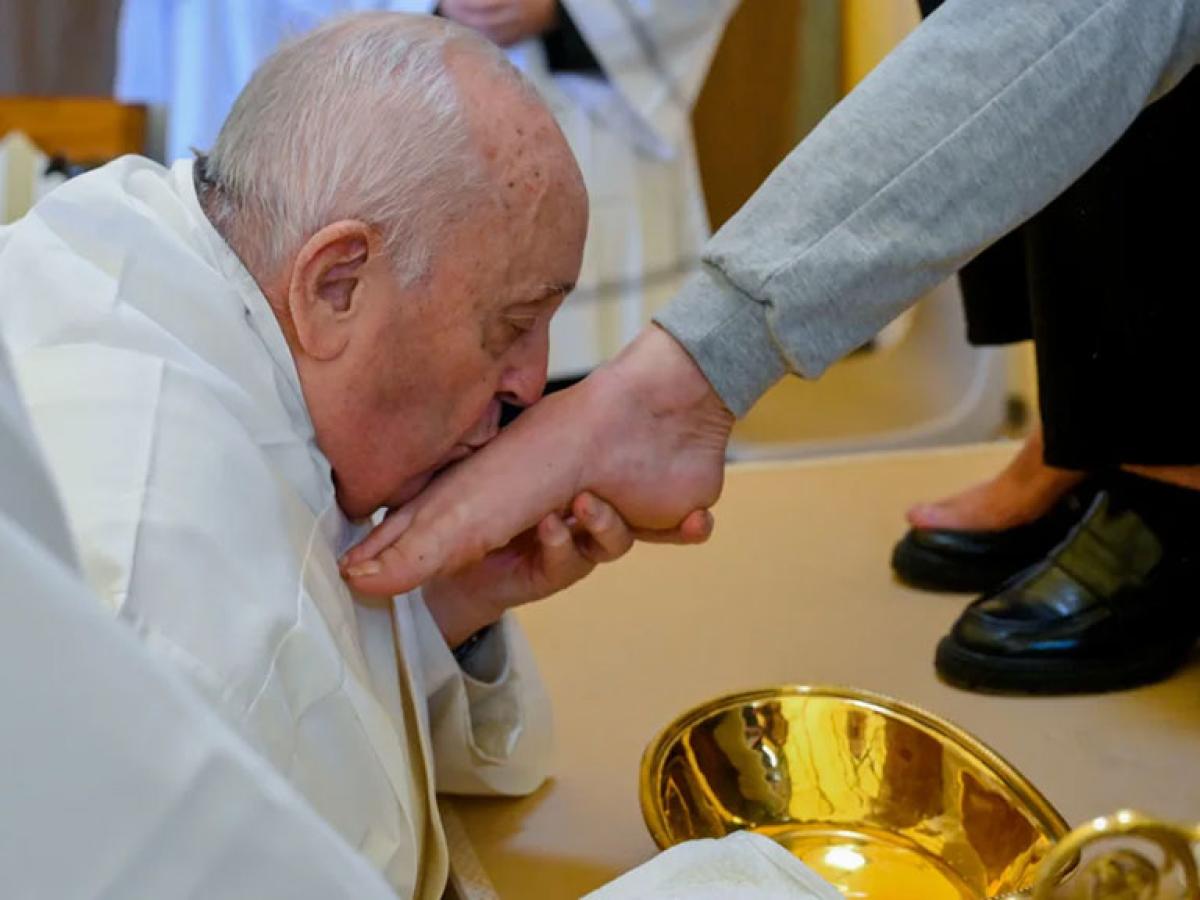 The Pope, wearing white, kneels, crades a bare foot, and kisses it.