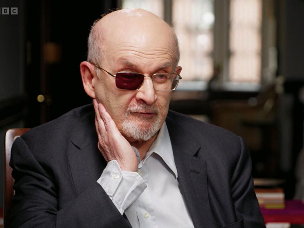 A man sits being interviewed and holds a hand to the side of his face, one lens of his glasses is tinted black.