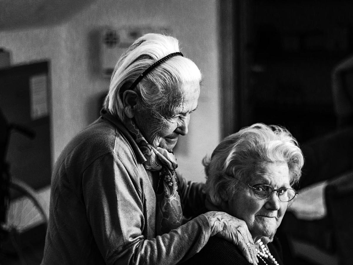A elderly women in a care home stands and places her hands on the shoulders of a seated woman.