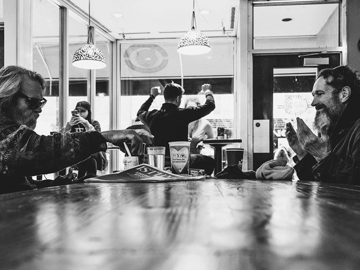 In a coffee shop, people sit around a table, one holds their hands up in surprise.