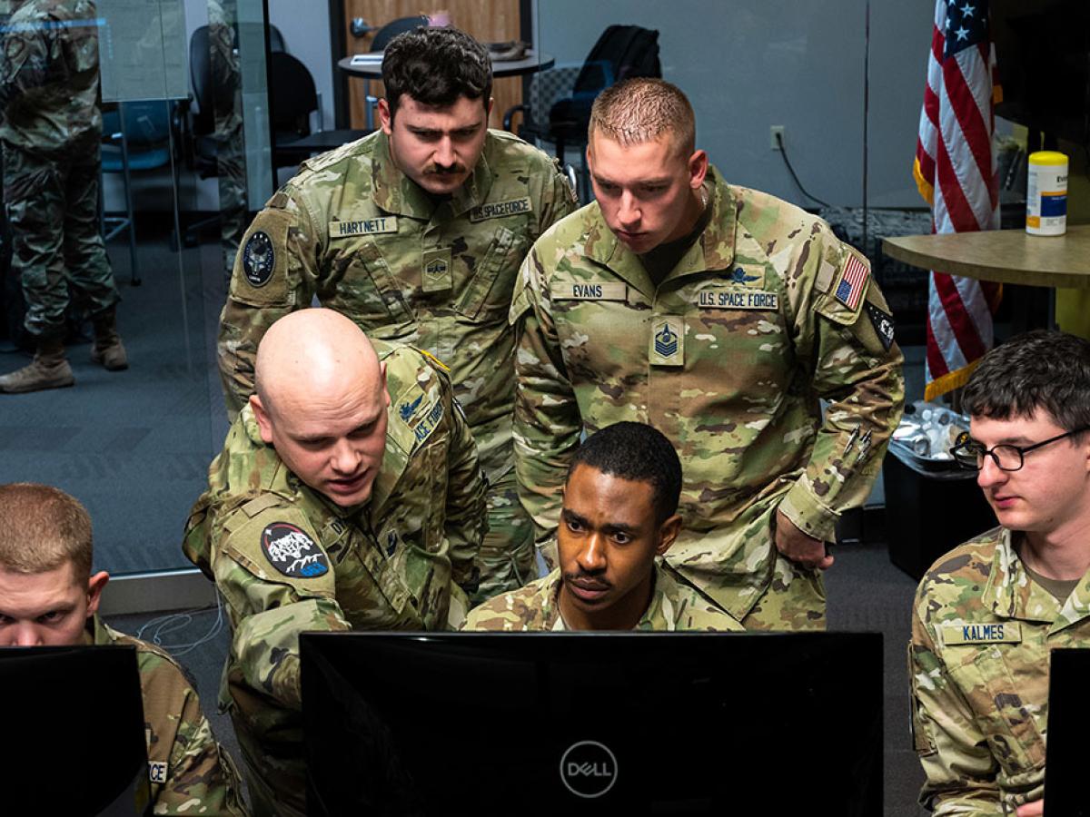 Military personnel, wearing camoflague uniform crowd round a computer monitor.