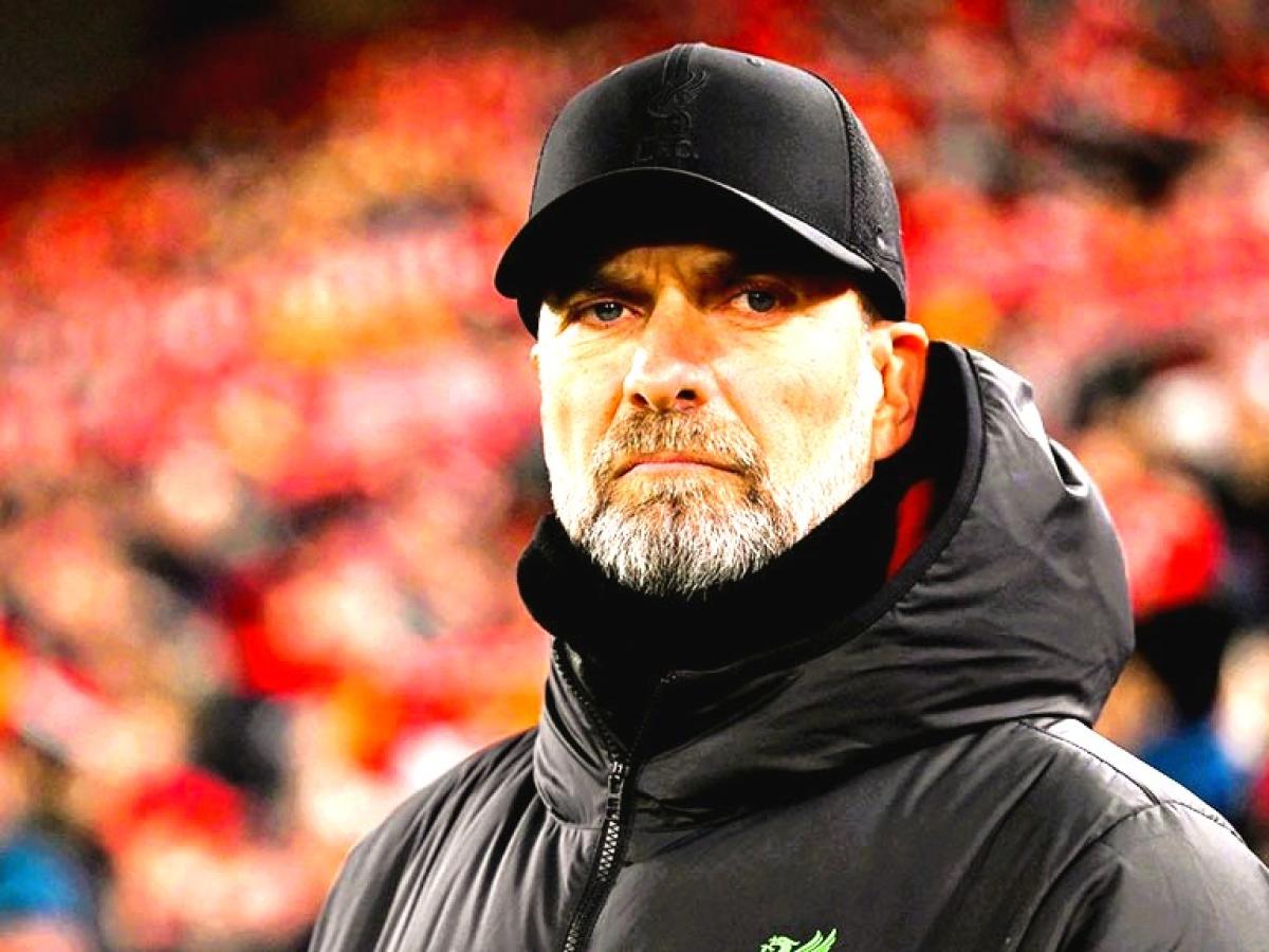 A stern looking football manager stares hard, with a stand of supporters behind him.