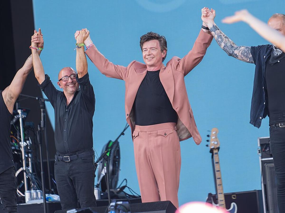 On a music festival stage, a popstar in a pink stages holds raised hands with his band.
