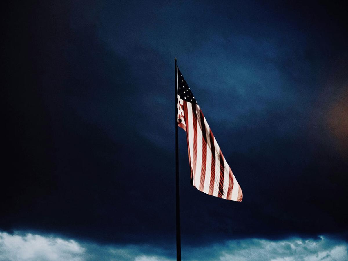 A US flag flutters under a dark brooding cloudy sky.