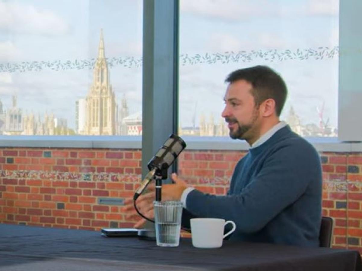 A podcast guest sits at a table talking with gesturing with their hands.