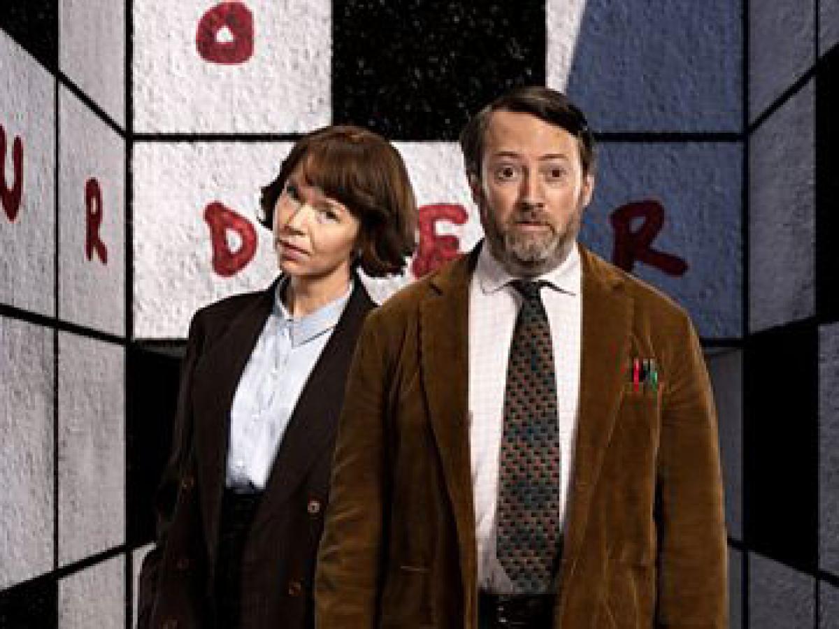 Two TV characters, a man and a woman, stand in front of a crossword cover walls of a room.