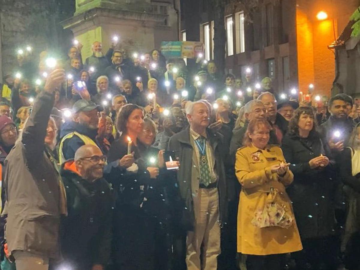 An outdoor vigil is lit by people holding up mobile phone lights.