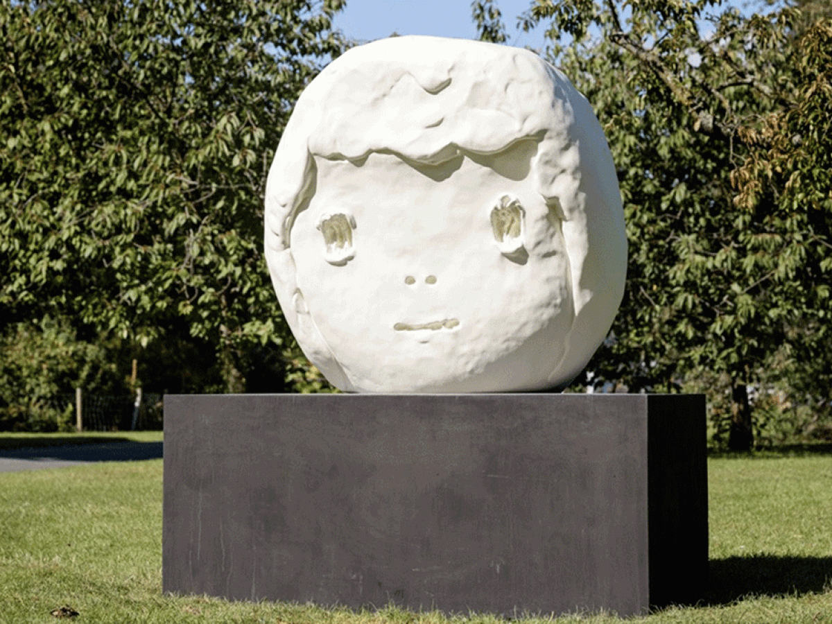 A large outdoor sculpture of a round child-like head sits on a dark plinth.