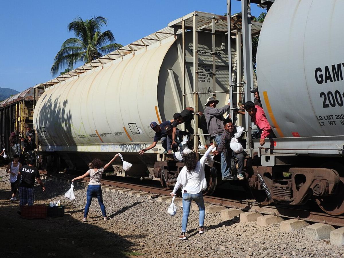 Migrants on a freight train reach for food bags held aloft by people on the track side.