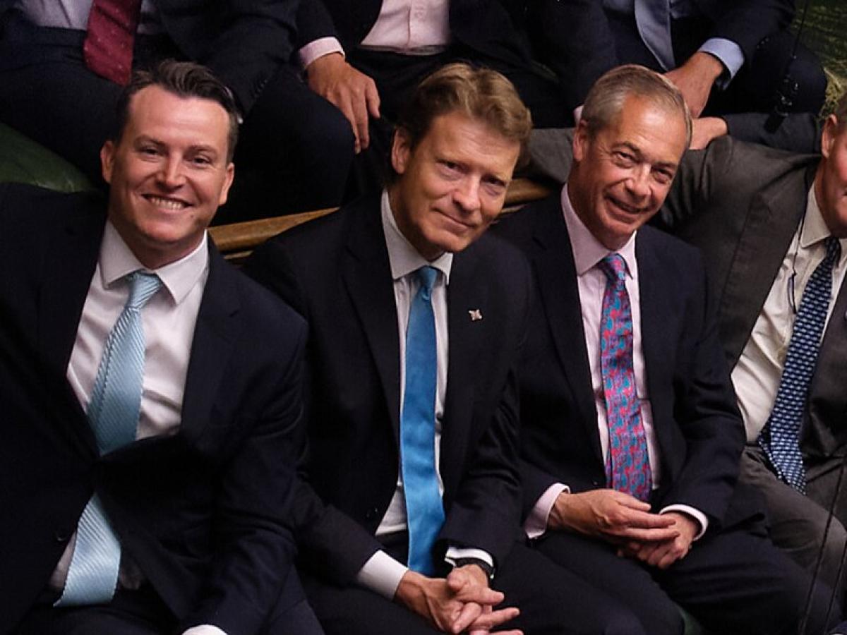 Four men in suits, sit next to each other smiling, in the House of Commons.