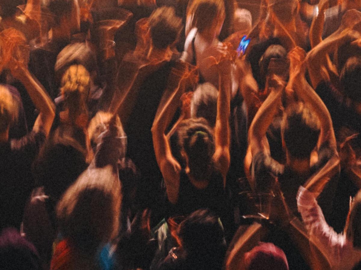 Concert goes stand, bathed in warm light, raising their hands in the air, facing away from the camera to the stage
