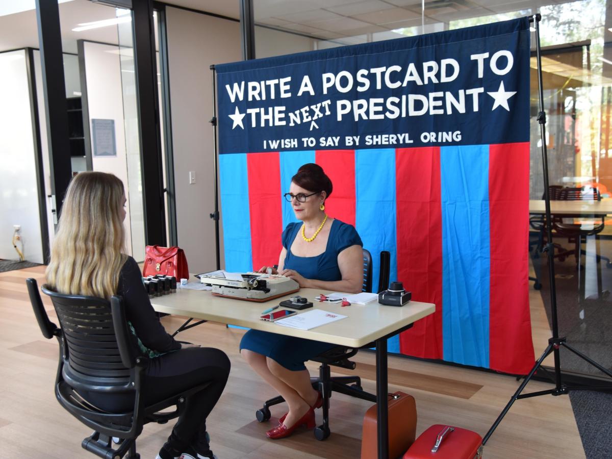 Two people sit at the table, one dictates as the other types. Behind a banner reads: write a postcard to the next president.i