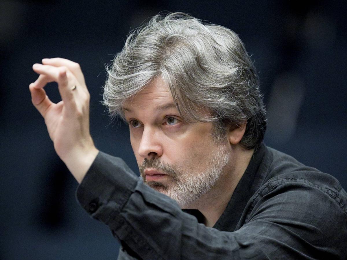 A conductor leans in toward an unseen orchestra with a raised hand.