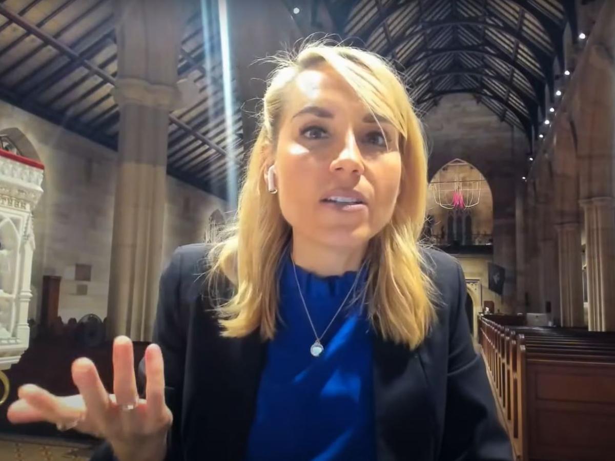 A woman sitting in an empty church talks to the camera and gestures with one hand.