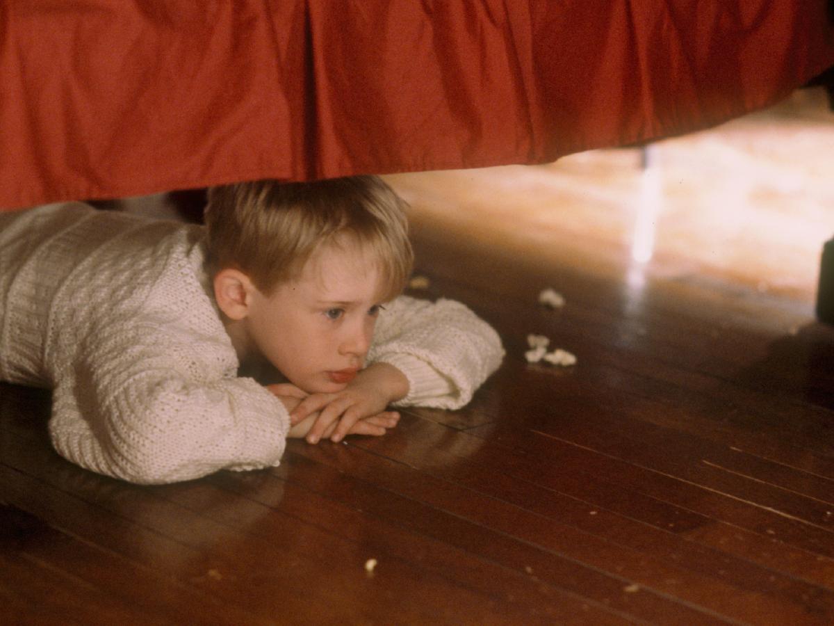 A child lies under a bed, resting his chin on his hands, contemplating.