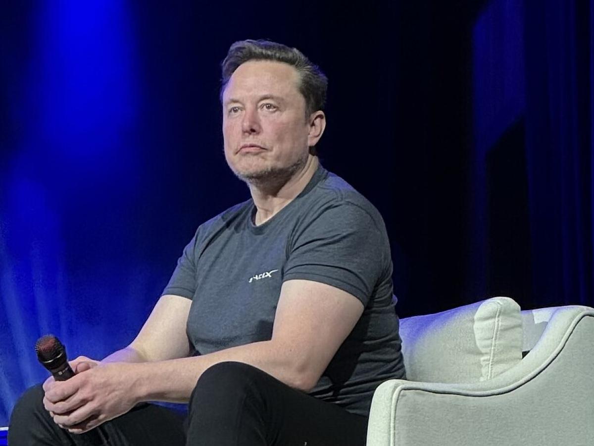 Elon Musk, wearing a t-shirt slouches forward, holding a mic, while sitting on a stage chair.
