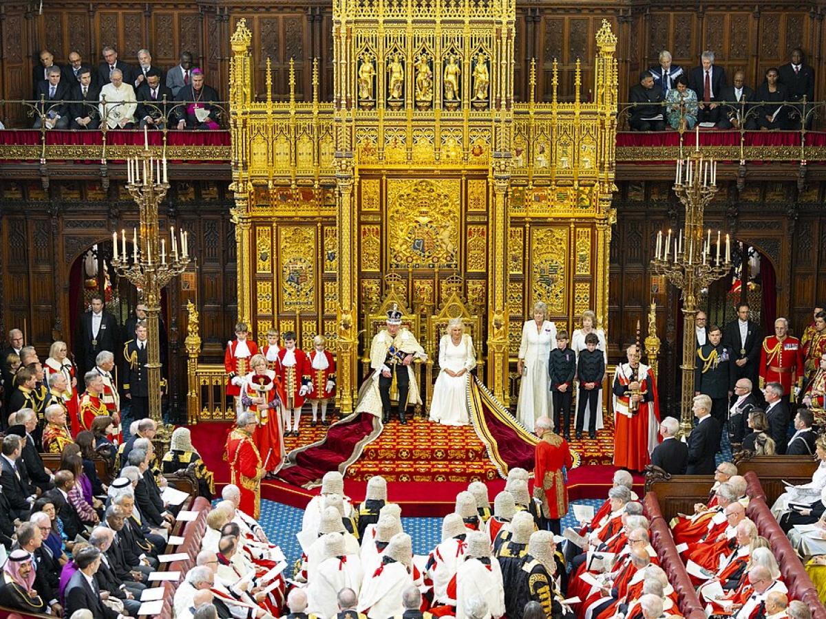 A wide angle picture shows a king and queen on thrones before many people in ceremonial clothes.