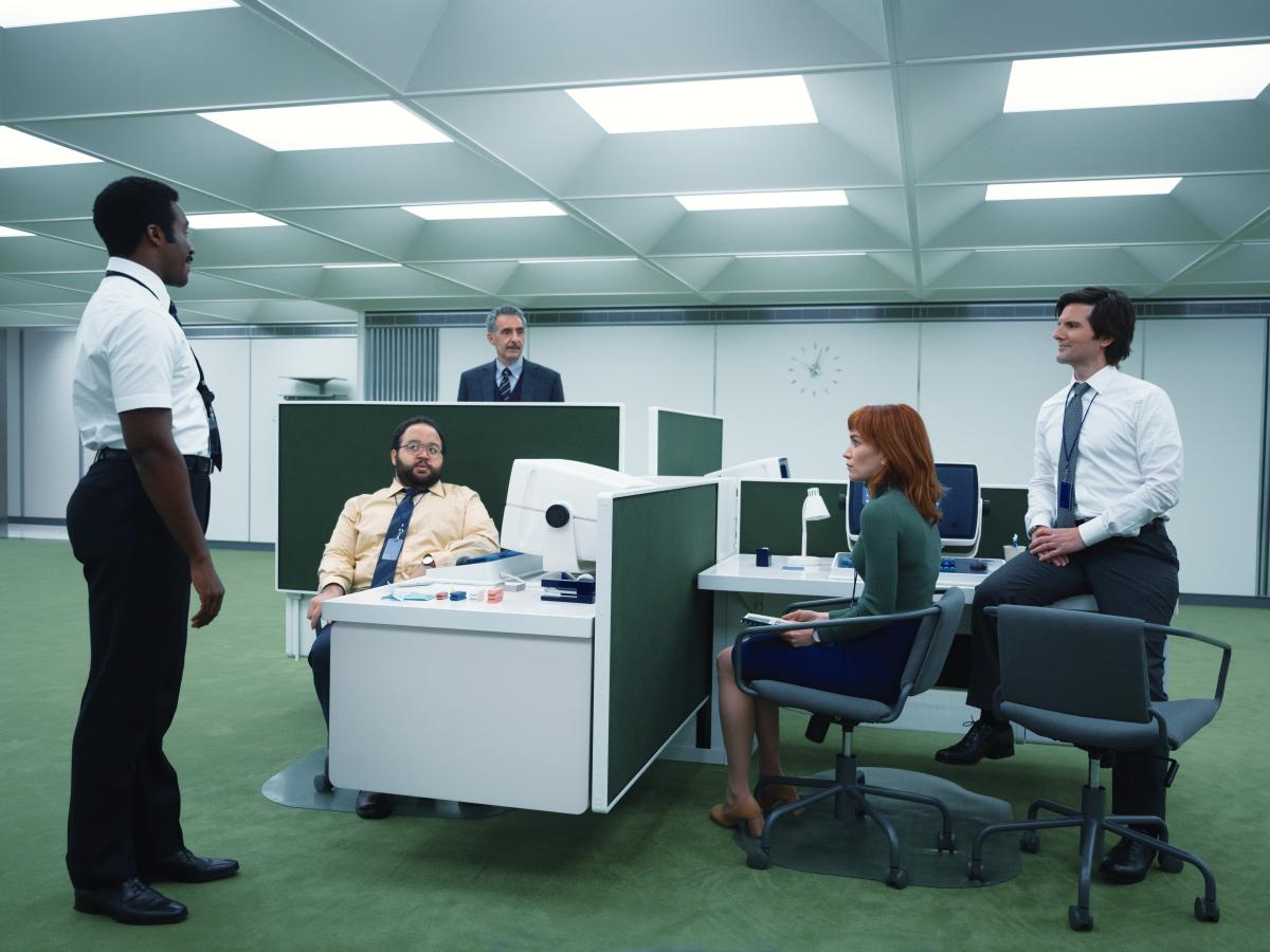 In a retro-future styled office, workers stand or sit around a pod of desks.