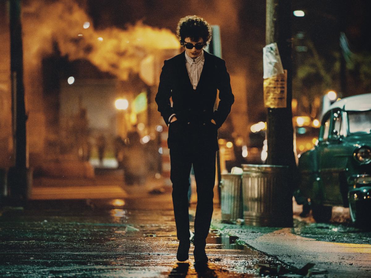 A hunched man wearing dark glasses and a dark suit, walks down a New York Street at night, with his hands in his pockets.