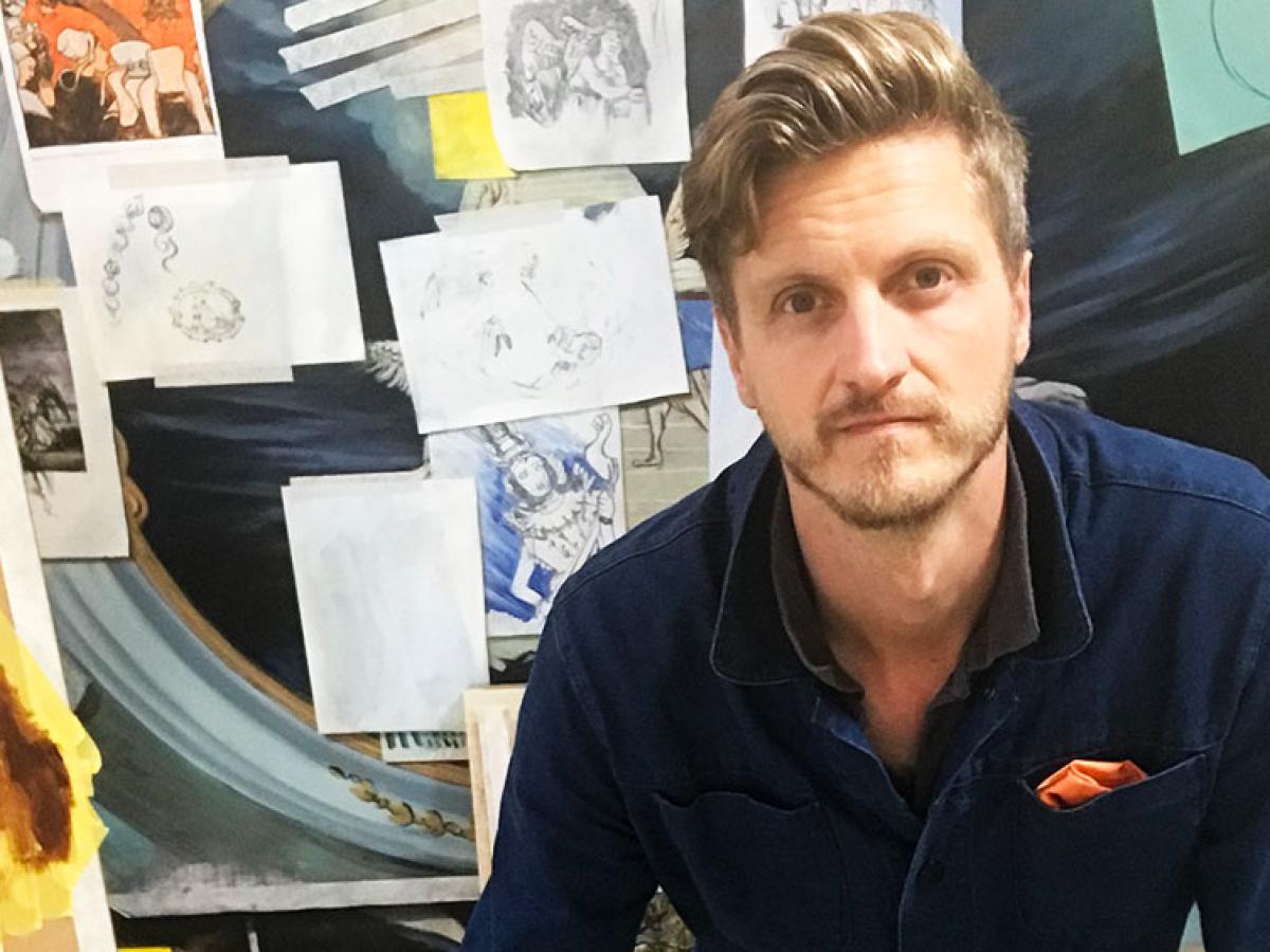 An artist sits in front of a board covered in images, canvases and paper.