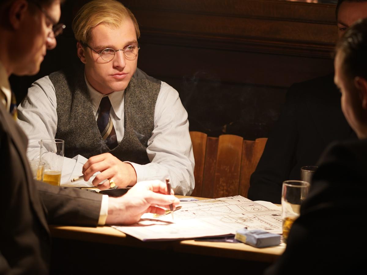 A man dressed in 1930s clothing, sits with others at a table looking pensive.