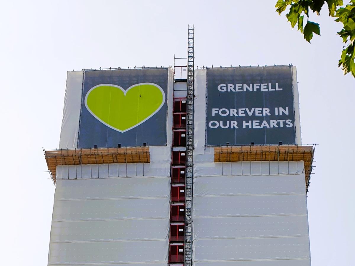 A white building wrap around a tower is topped by a green heart and the slogan: Grenfell Always In Our Heaets.
