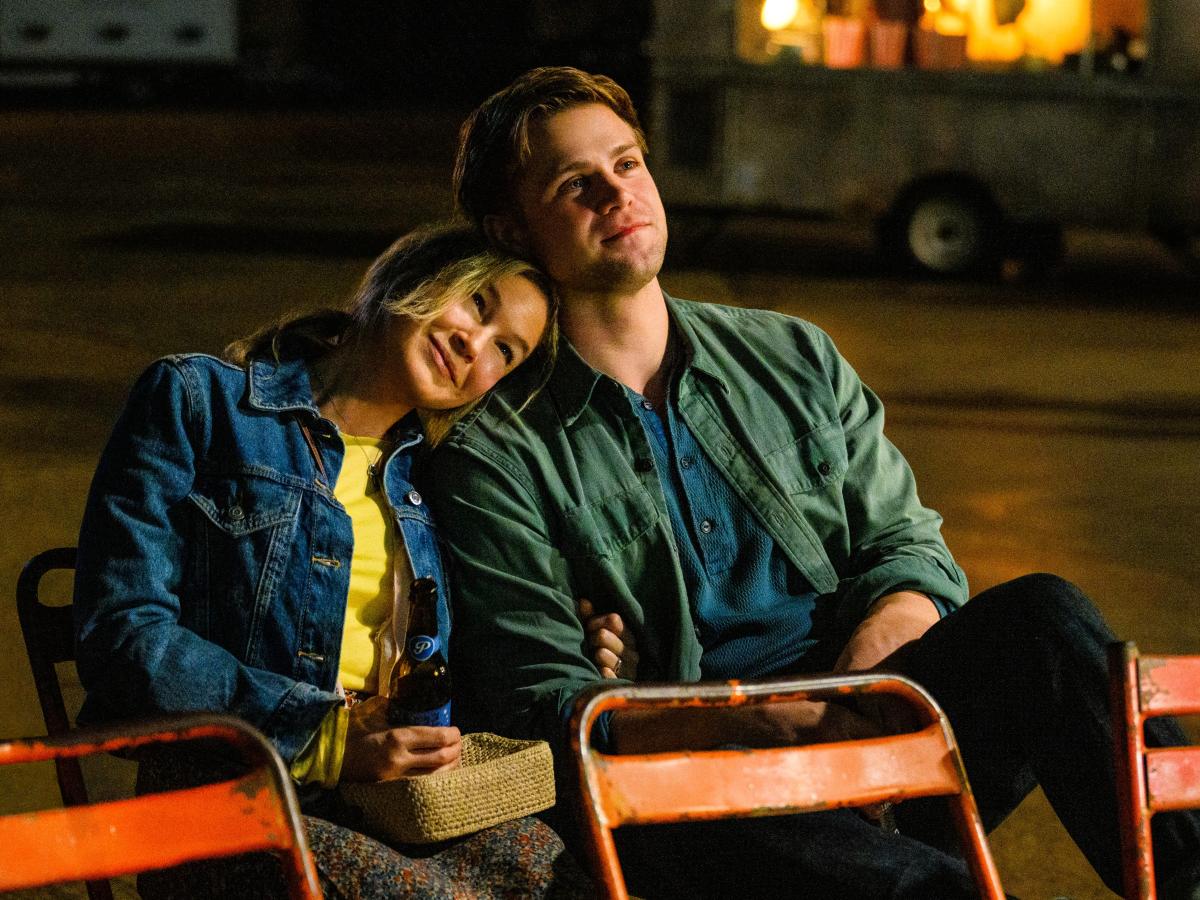 A couple sit on outdoor seats, her resting her head on his shoulder.