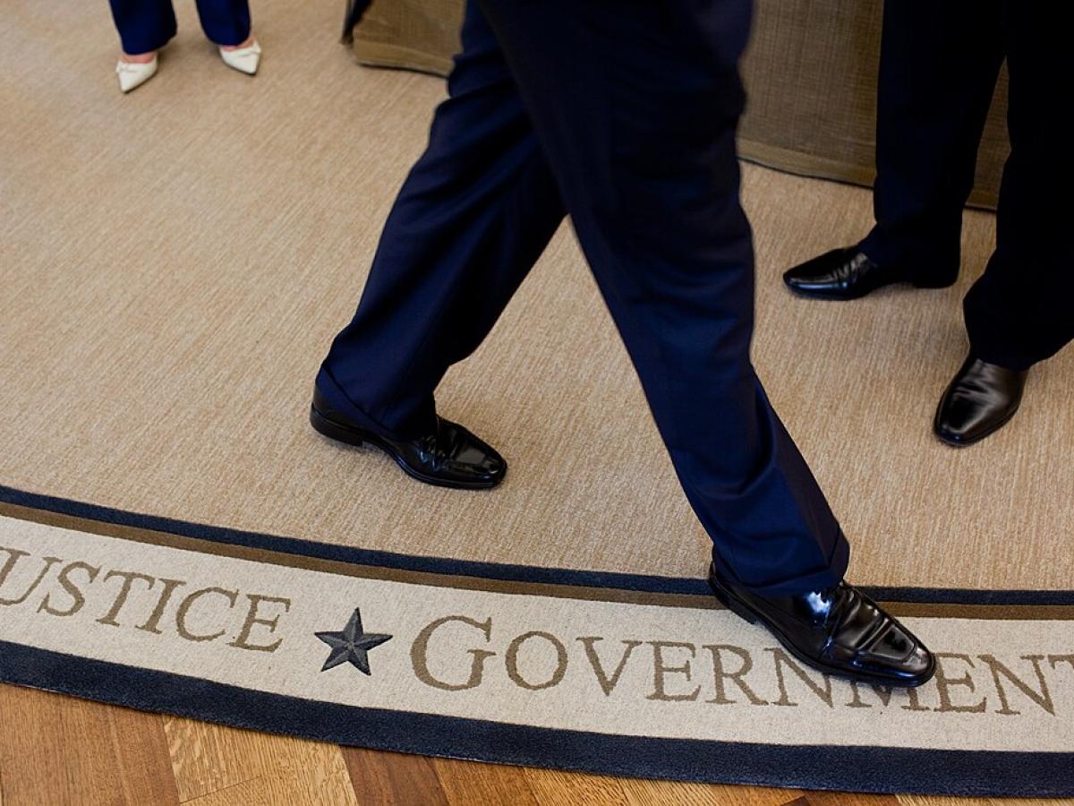 The feet and legs of someone walking on a white rug, beside the words Justice and Government woven in to it,