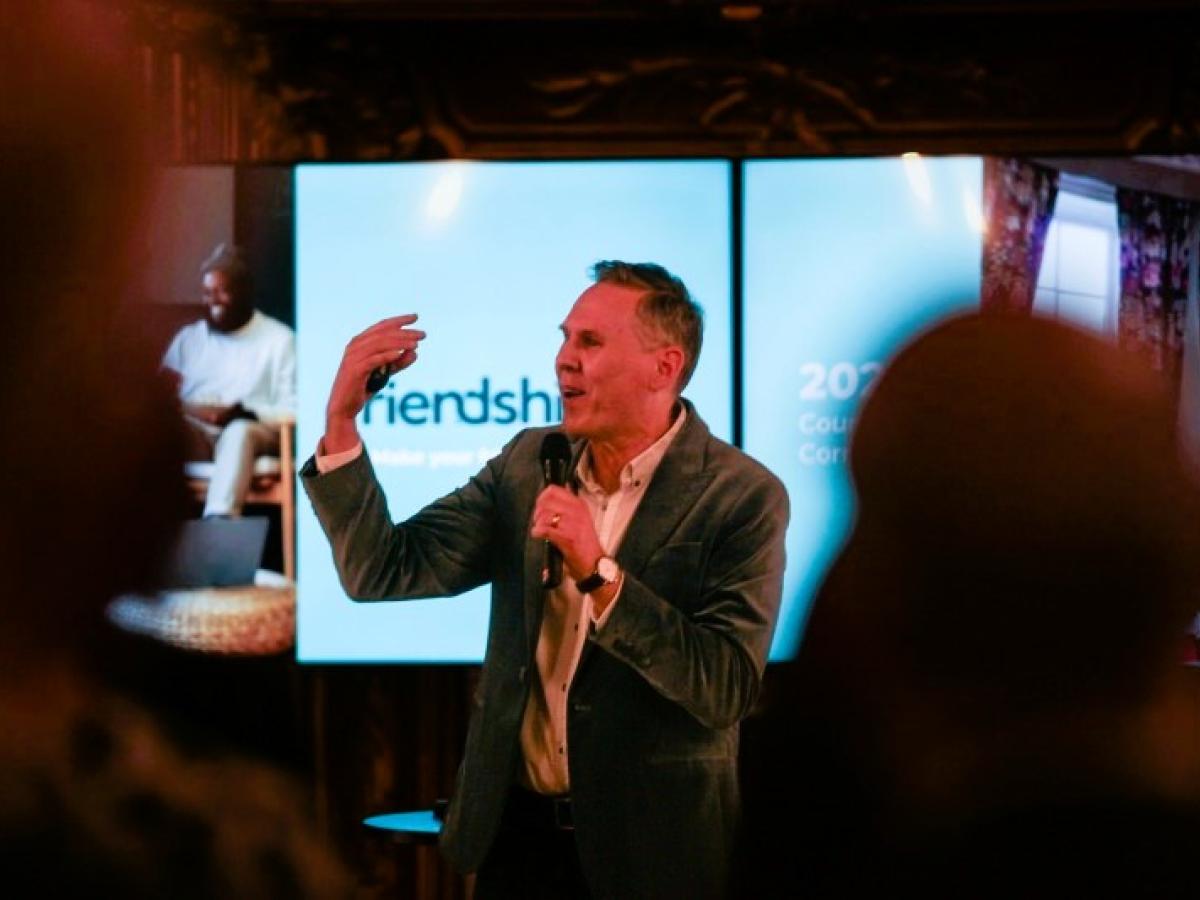 A speaker, standing in front of a screen, beckons with one hand, holding a mic with the other.