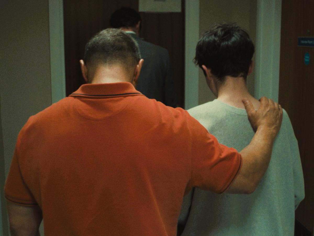   A father walks with his son away from the camera, his hand on his son's shoulder.