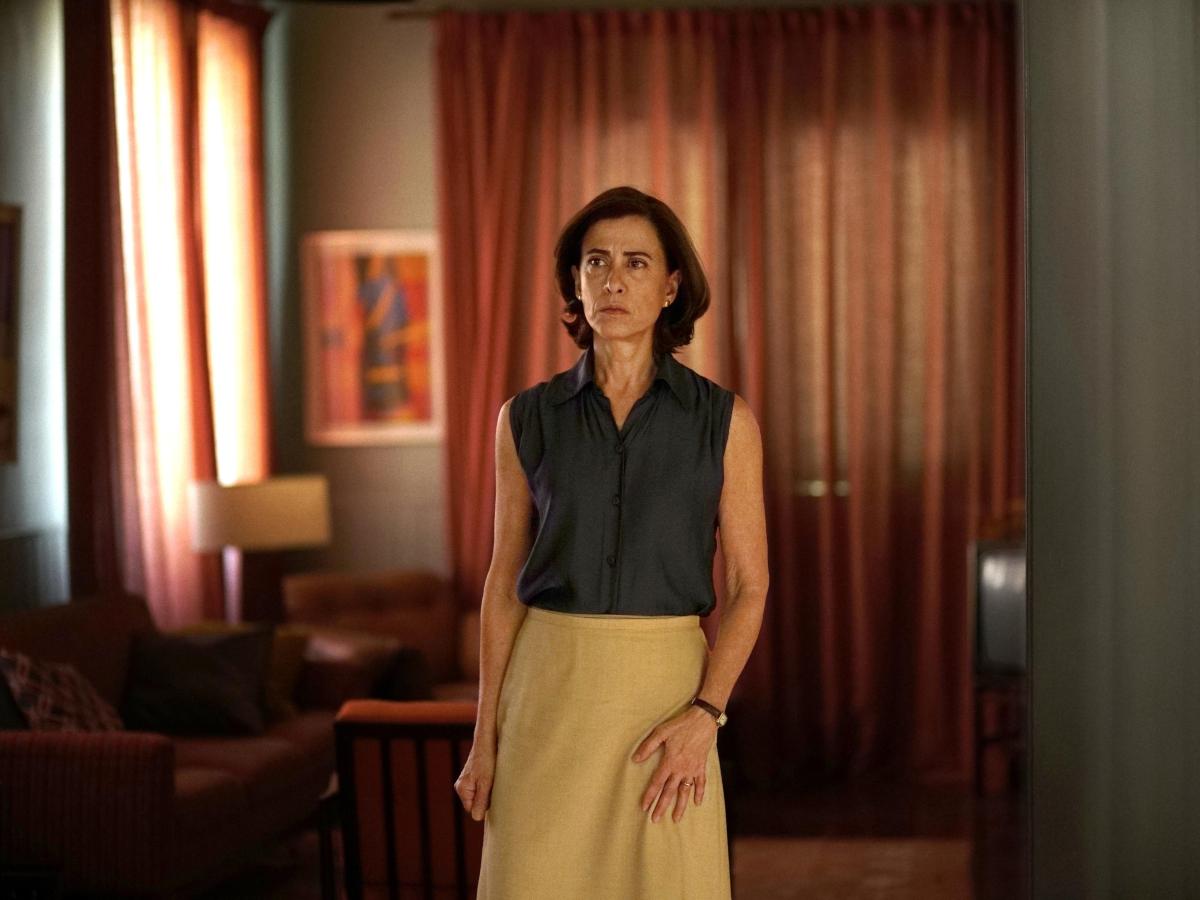A woman, wearing 70s clothing, stands, looking apprehensive.